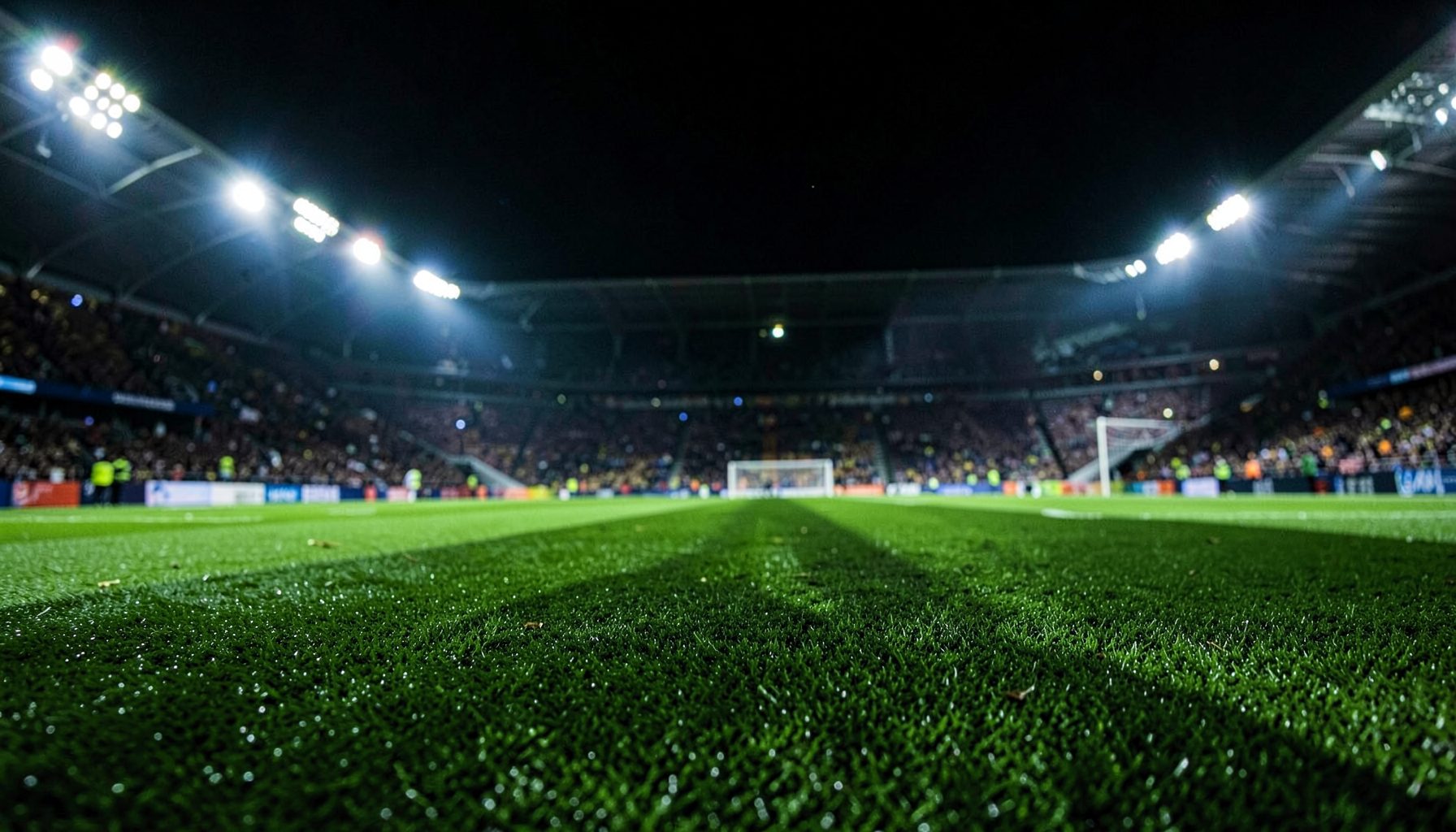 Close-up of a football pitch under stadium floodlights at night with deep green turf and dramatic shadows