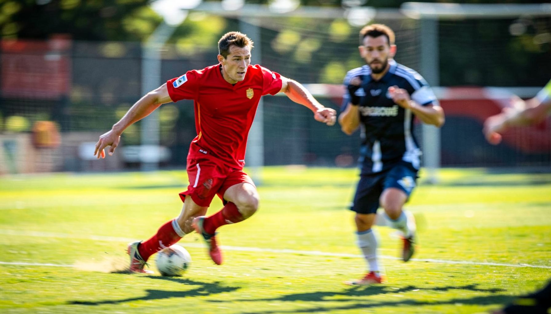A player in a red jersey dribbling past a defender on a sunlit pitch, motion blur conveying speed and intensity