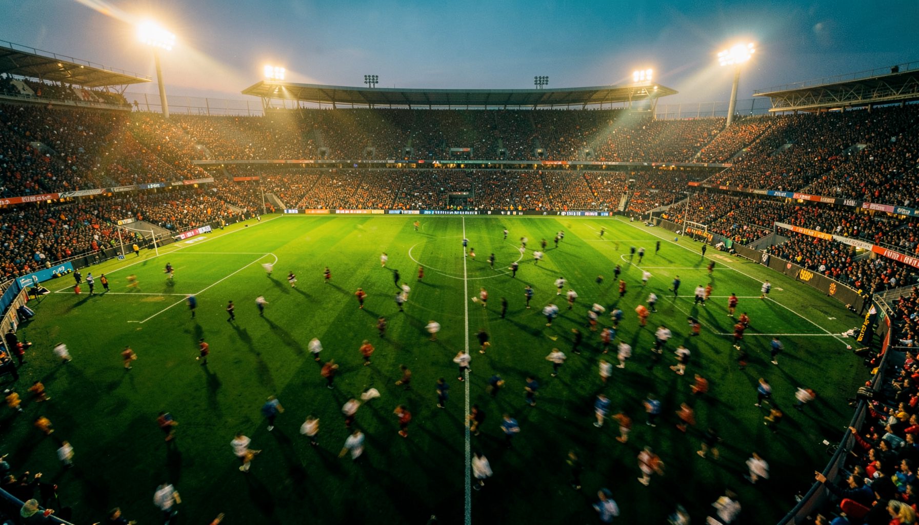 A wide aerial view of a packed football stadium at dusk under floodlights, deep green turf glowing, crowd blurred in motion
