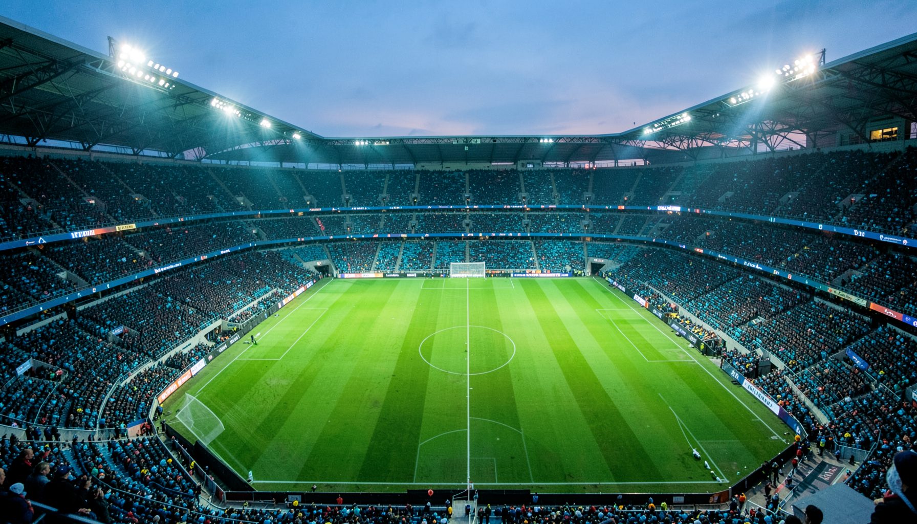 Wide aerial view of a packed football stadium at dusk with floodlights blazing across a green pitch in deep teal and navy tones