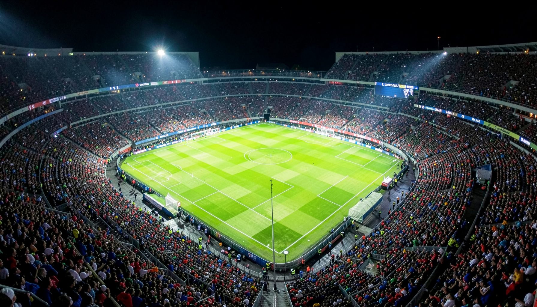 Aerial view of a Champions League stadium at night, the pitch glowing under bright lights with a sea of fans in the stands